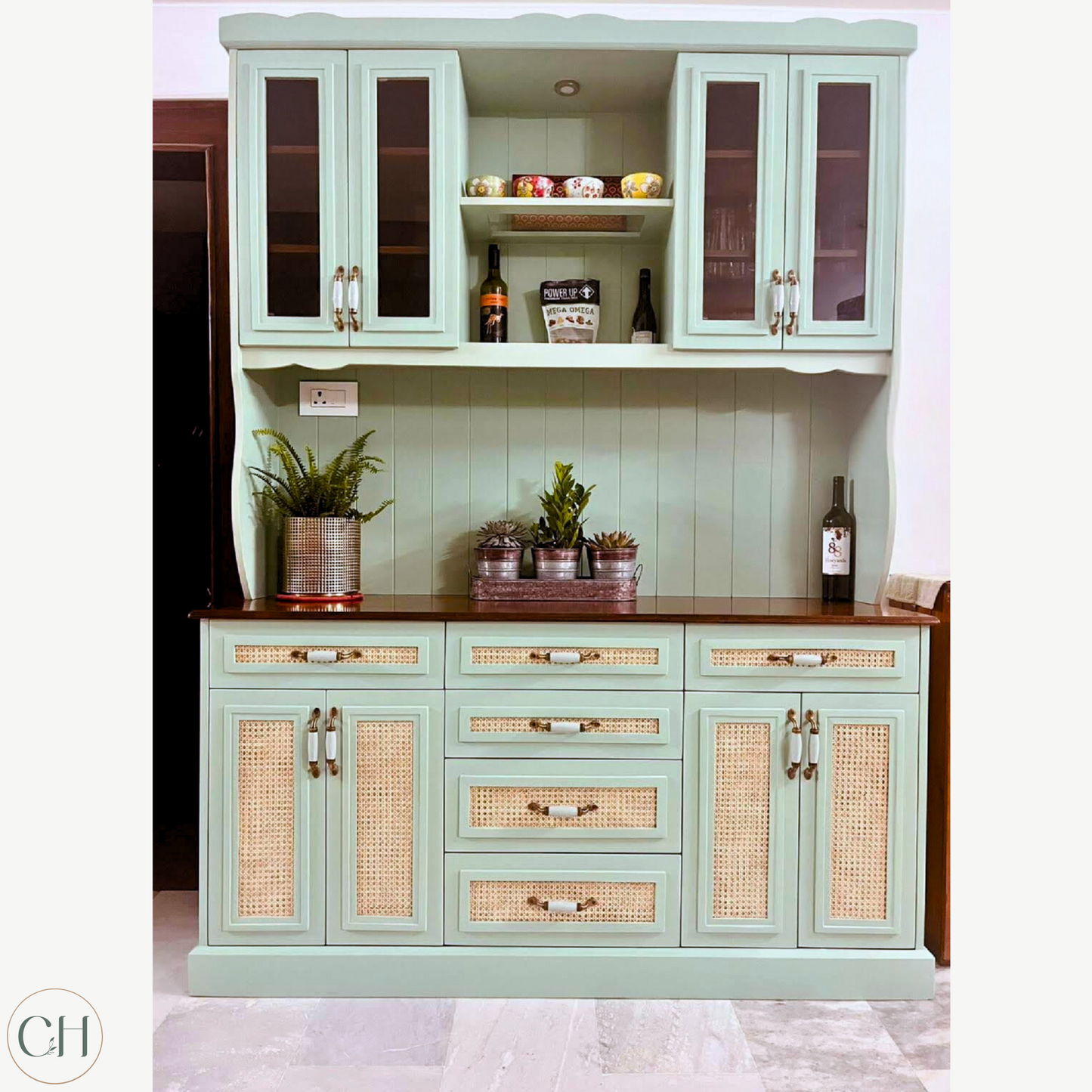 CustHum - Large farmhouse-style teal-colored crockery cabinet hutch in birch ply with rattan and glass panels and modern enamel handles, displaying crockery and planters (front view).