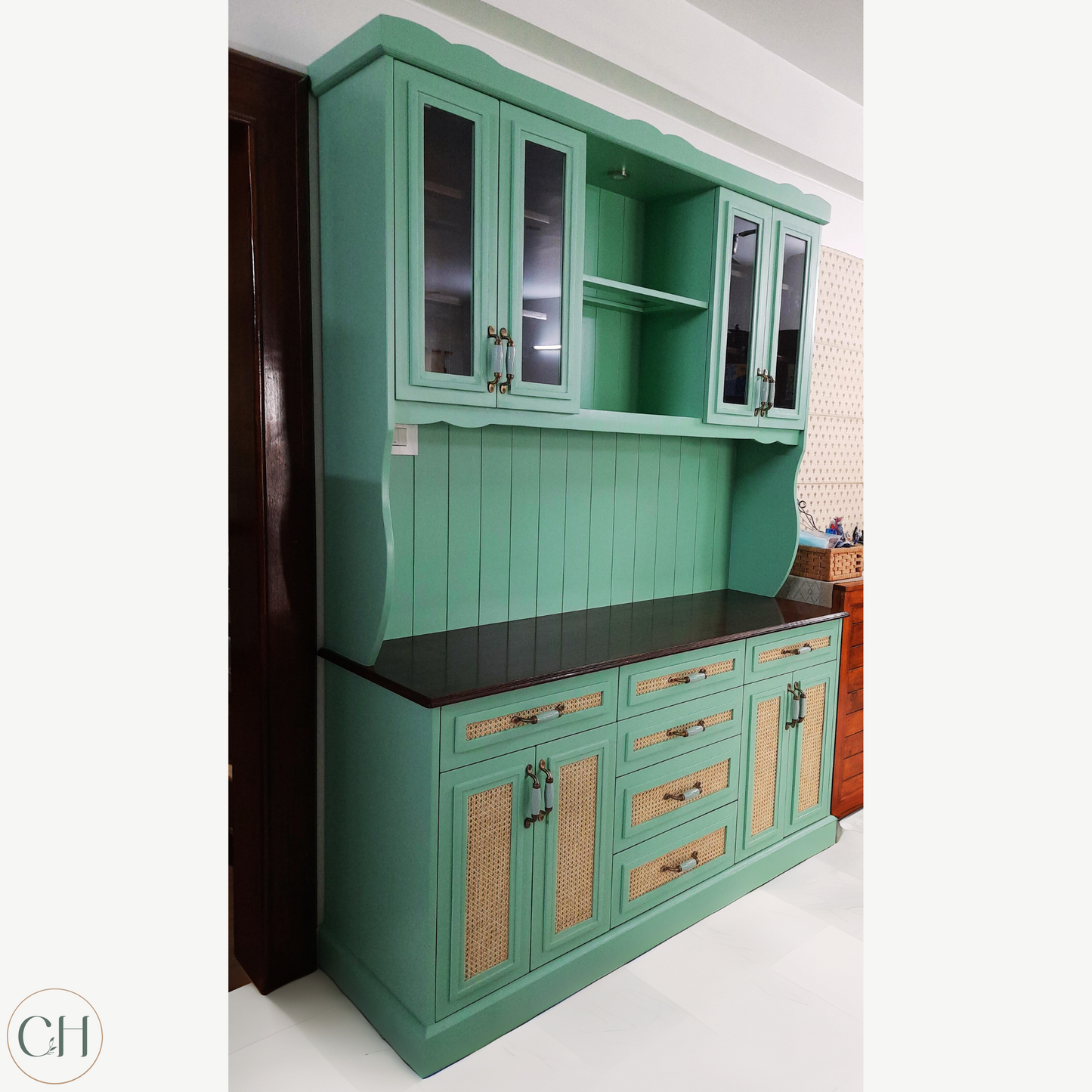 CustHum - Large farmhouse-style teal-colored crockery cabinet hutch in birch ply with rattan and glass panels and modern enamel handles, displaying crockery and planters (ISO view).