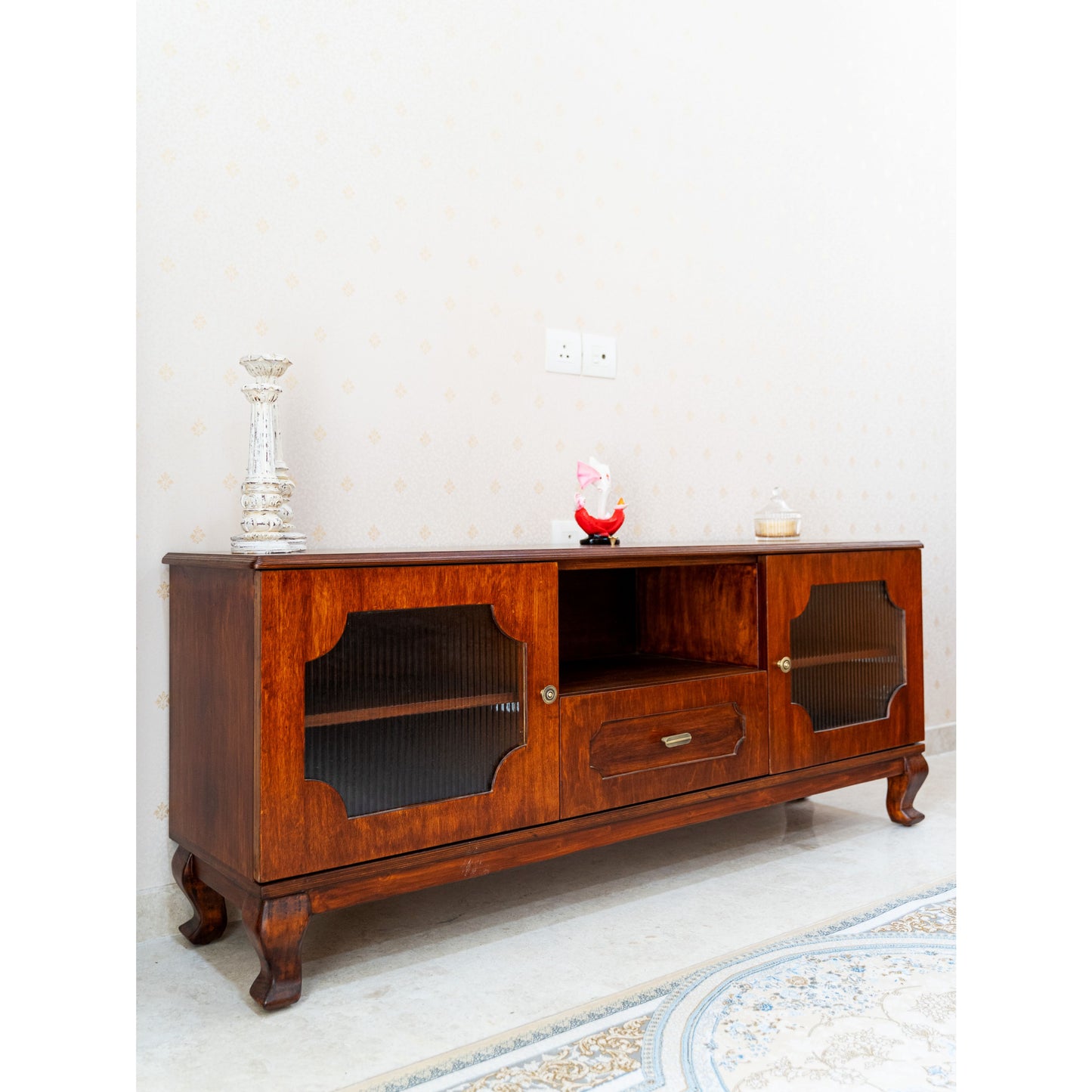 CustHum - ISO view of an ornate wooden TV stand with cabriole legs, fluted-glass door panels, open shelf and drawer in the middle; styled with small objets d'art